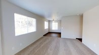 This single-floor residence offers a clean, open layout designed for comfortable everyday living. The main living area is generously proportioned, featuring plush wall-to-wall carpeting that transitions into hard-surface flooring in the adjoining space, creating a natural visual distinction between living and dining or entryway zones. Neutral, light-toned walls throughout lend the interior a bright, airy quality that enhances the sense of openness. Multiple windows along the exterior wall flood the space with abundant natural light, offering views of the surrounding greenery outside. A ceiling fan centered overhead in the adjoining area provides both functional air circulation and a subtle design accent. The smooth white ceiling and consistent neutral color palette create a cohesive, understated aesthetic throughout. The layout flows intuitively from one area to the next, making the space feel connected and well-organized. Baseboards and clean wall finishes reflect a well-maintained interior. Overall, the residence presents a straightforward, functional design that balances practicality with a welcoming, comfortable ambiance suitable for a variety of living arrangements.