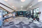 Located at 525 North Ave NE in Atlanta's Old Fourth Ward neighborhood, this fitness center is a well-appointed, single-level facility designed to support a comprehensive workout experience. The space is defined by its open, elongated layout, which creates a sense of generous floor space and allows for clear separation between functional training zones.

The flooring throughout features a distinctive geometric hexagonal carpet tile pattern in varying shades of grey, adding a modern, design-forward aesthetic. Pendant lighting and recessed ceiling fixtures provide ample illumination, while large floor-to-ceiling windows and glass doors flood the space with natural light and offer views of an adjacent outdoor patio area, which is accessible directly from the fitness center.

The cardio zone is equipped with treadmills, stationary cycling bikes, and a stair climber, many positioned to face mounted flat-screen televisions. The strength training area includes a cable machine system, a TRX suspension training rig, free weights, a weight bench, and plyo boxes for functional training. Full-length mirrors with teal accent wall panels and bold diagonal trim details enhance the space visually while supporting form-checking during workouts.

Overall, the fitness center presents a clean, contemporary ambiance that balances athletic functionality with polished interior design.