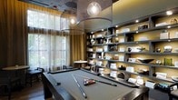 This single-floor space is designed as a sophisticated game and lounge area, combining functionality with contemporary interior design. The centerpiece of the room is a full-size billiard table finished in a sleek gray felt, positioned beneath two large, industrial-style mesh drum pendant lights that cast a warm, focused glow over the playing surface. The lighting fixtures serve as strong architectural statements, complementing the room's modern aesthetic.

Floor-to-ceiling open shelving lines one wall, featuring illuminated compartments that display an artfully curated collection of decorative objects, books, and sculptural pieces in neutral tones. The shelving system is framed in dark metal, lending a structured yet refined look to the space.

Large windows draped with sheer linen curtains allow abundant natural light to filter into the room, offering views of lush greenery outside. A small round table with seating is positioned near the windows, providing an intimate conversation or casual lounge area.

The overall ambiance is upscale and inviting, with a layered design palette of warm golds, deep grays, and natural textures that create a cohesive, club-like atmosphere suited for both leisure and socializing.