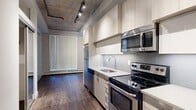 Located on the sixth floor at 10620 116 Street NW in Edmonton, this one-bedroom, one-bathroom unit showcases a compelling blend of industrial and contemporary design. The exposed concrete ceiling with visible mechanical elements and track lighting lends the space an urban loft aesthetic, while warm wood-tone vinyl plank flooring runs throughout, creating a cohesive and inviting atmosphere.

The galley-style kitchen is well-appointed with light wood cabinetry extending to the ceiling, sleek white countertops, a stainless steel undermount sink, and a full suite of stainless steel appliances including an electric range, built-in microwave, and refrigerator. The open sightline from the kitchen toward the living area emphasizes the efficient, thoughtfully organized layout.

The bathroom features a modern vanity with a stone-topped sink, a mirrored medicine cabinet, and a glass-enclosed shower with tiled surround. The bedroom benefits from natural light through a large window and includes a ceiling fan, with an adjacent walk-in closet providing ample storage.

A standout convenience is the in-suite stacked washer and dryer, neatly tucked into a dedicated closet. Throughout the unit, clean white walls and carefully considered finishes contribute to a bright, polished feel that balances functional urban living with stylish, modern design.