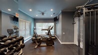 Located at 2701 E Lookout Dr in Garland, TX, this single-floor fitness center is a comprehensively equipped exercise facility designed to support a wide range of workout routines. The room features a two-toned color scheme of teal and gray walls, accented by white crown molding and recessed lighting throughout, creating a clean and energizing atmosphere. Rubber flooring provides durability and safety underfoot across the entire space.

The cardio section is outfitted with multiple Cybex machines, including stationary bikes and an elliptical trainer, positioned near large windows with slatted blinds that allow ample natural light while maintaining privacy. A wall-mounted flat-screen television is positioned for easy viewing during workouts.

The strength training area includes a full dumbbell rack with an extensive range of weights, complemented by a large wall mirror that enhances the sense of space and allows users to monitor their form. A multi-functional cable machine with various attachments supports a broad spectrum of resistance exercises, while TRX suspension straps add versatility. Resistance bands and a stability ball, accompanied by posted exercise guides, round out the functional training options.

Ceiling fans provide additional air circulation, and a water cooler ensures hydration is always within reach. The space is well-organized and thoughtfully arranged to maximize usability for all fitness levels.
