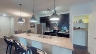 This single-floor residence showcases a thoughtfully designed open-concept layout that seamlessly connects the kitchen and dining areas, creating an inviting and functional living environment. The kitchen is a standout feature, boasting rich dark cabinetry that contrasts beautifully with the light granite countertops. A large kitchen island with an undermount stainless steel sink serves as both a functional workspace and a casual dining spot, complete with bar seating. Industrial-style pendant lights hang overhead, casting a warm, focused glow across the space and adding a contemporary edge to the design. The full suite of stainless steel appliances — including a French door refrigerator, gas range, and built-in microwave — offers both style and practicality. A mosaic tile backsplash adds texture and visual interest to the cooking area, complementing the overall modern aesthetic. A dedicated wine rack built into the upper cabinetry provides a refined touch for entertaining. The adjacent dining area is visible from the kitchen, reinforcing the open and connected feel of the floor plan. The overall ambiance is modern and polished, with cohesive design choices that create a sleek, well-appointed living space.