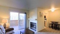 This single-floor residence offers a thoughtfully arranged layout with a comfortable, functional design. The living area serves as the central gathering space, anchored by a fireplace with a tiled surround, a classic white mantel, and gold-accented glass doors, making it a natural focal point of the room. Built-in wall shelving flanks the fireplace, providing convenient display or storage space. The living area is finished with wall-to-wall carpeting, contributing to a warm and cozy atmosphere.

A large sliding glass door fills the space with natural light and opens directly onto a private outdoor balcony enclosed by a wooden railing, offering views of mature evergreen trees and the surrounding neighborhood. The seamless indoor-outdoor connection enhances the sense of openness in the living area.

Adjacent to the living space, a glimpse into the kitchen reveals white cabinetry, standard appliances including a microwave and stove, and a practical, compact layout. The overall interior features neutral walls and light-toned ceilings, creating a bright and airy ambiance throughout the home.