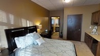 This single-floor space offers a thoughtfully designed layout centered around a cozy and stylish bedroom. The room is finished with warm, earthy brown-toned walls that create an inviting and relaxing ambiance. A drop tile ceiling with recessed and flush-mount lighting provides even, comfortable illumination throughout the space. The bedroom is furnished with a generously sized bed dressed in elegant, patterned bedding accented with decorative pillows, complemented by a sleek dark-wood headboard. A matching dark-wood desk and chair sit beside the bed, offering a functional workspace. A tall decorative floor vase with dried branches adds a refined, organic touch to the room's aesthetic. Rich, dark-toned interior doors further enhance the space's sophisticated color palette. Through the open doorway, an en-suite bathroom is visible, featuring warm lighting and tiled flooring, providing convenient private access directly from the bedroom. The neutral carpeted flooring ties the room together cohesively. Overall, the space delivers a comfortable and polished environment with a consistent design language of dark wood accents, warm neutral tones, and practical amenities.