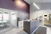 The Edge at Greentree is a professionally designed fitness and wellness facility located at 1000 Cedartree Lane in Claymont, Delaware. The single-floor space makes efficient use of its footprint, offering a clean, contemporary aesthetic throughout. Guests are welcomed by a sleek reception area anchored by a wood-paneled front desk set against a bold purple textured accent wall bearing the facility's branding. The lobby features large-format tile flooring, soaring vaulted ceilings, track lighting, and a comfortable seating area bathed in natural light from windows overlooking the exterior. The expansive exercise room is the centerpiece of the facility, equipped with a wide array of cardio machines, cable and strength training systems, a dumbbell rack, medicine balls, resistance equipment, and floor-to-ceiling mirrors that amplify the sense of space. Distinctive purple overhead ductwork adds an industrial design accent against the bright white walls and vaulted ceiling. A dedicated meeting and dining room provides an intimate, well-appointed space with a long white conference table, mesh seating, built-in cabinetry, decorative wall graphics, and natural light from roller-shade windows. The hallway connects each area seamlessly, maintaining the facility's cohesive modern and professional atmosphere throughout.