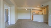 This single-floor space showcases an open-concept design that seamlessly connects the living area to the kitchen. The living space is finished with neutral wall-to-wall carpeting, creating a warm and understated atmosphere. Walls are painted in a soft, neutral tone throughout, lending a clean and cohesive look to the interior.

A notable design feature is the pendant light fixture suspended from the ceiling, which serves as a visual anchor between the living and dining areas, adding a subtle decorative touch. The ceiling height provides a comfortable sense of openness in the main living area.

The kitchen is positioned at the rear of the open layout and is defined by light wood cabinetry, white appliances including a microwave and range, and a raised breakfast bar that doubles as a partition between the kitchen and the living space. A fluorescent ceiling fixture provides direct task lighting over the kitchen area. The breakfast bar offers casual seating potential and enhances the functional flow of the space. Overall, the layout is practical and well-suited for everyday living.