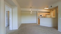 This single-floor space showcases an open-concept design that seamlessly connects the living area to the kitchen. The living space is finished with neutral wall-to-wall carpeting, creating a warm and understated atmosphere. Walls are painted in a soft, neutral tone throughout, lending a clean and cohesive look to the interior.

A notable design feature is the pendant light fixture suspended from the ceiling, which serves as a visual anchor between the living and dining areas, adding a subtle decorative touch. The ceiling height provides a comfortable sense of openness in the main living area.

The kitchen is positioned at the rear of the open layout and is defined by light wood cabinetry, white appliances including a microwave and range, and a raised breakfast bar that doubles as a partition between the kitchen and the living space. A fluorescent ceiling fixture provides direct task lighting over the kitchen area. The breakfast bar offers casual seating potential and enhances the functional flow of the space. Overall, the layout is practical and well-suited for everyday living.