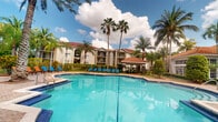 This single-story residence is part of a well-appointed tropical community defined by its Mediterranean-inspired architectural style. The buildings feature warm stucco exteriors, red clay tile rooflines, and private balconies that overlook the lushly landscaped grounds. Towering palm trees frame the property, creating a resort-like atmosphere that is characteristic of South Florida living.

The standout feature of the community is the expansive outdoor swimming pool, which serves as a central gathering point. The generously sized pool is surrounded by a spacious deck furnished with bright blue lounge chairs, providing ample room for sunbathing and relaxation. A covered cabana pavilion anchors the pool area, offering a shaded retreat with additional seating and amenities for residents. The surrounding landscaping is verdant and well-maintained, with dense tropical greenery, mature palms, and manicured shrubs that enhance the sense of privacy and seclusion.

The open-air setting benefits from abundant natural light throughout the day, and the overall ambiance evokes a laid-back, vacation-inspired lifestyle. The combination of classic architectural detailing and resort-style outdoor amenities makes this a distinctive and inviting community environment.