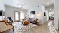 This single-story residence showcases a cohesive mid-century modern aesthetic carried throughout its well-designed layout. Light wood-style flooring flows continuously across the space, complementing the bright white walls and creating a warm, airy atmosphere.

The living area serves as the heart of the home, featuring a ceiling fan, sheer curtain-framed glass door that invites natural light, and direct outdoor access. The space comfortably accommodates a generous seating arrangement anchored by a geometric-patterned area rug.

The kitchen is thoughtfully designed with flat-front cabinetry in a warm wood tone, white quartz countertops, subway tile backsplash, and a full suite of stainless steel appliances. A peninsula with seating provides both additional prep space and casual dining.

Two bedrooms offer restful retreats, each featuring large windows with layered sheer and blackout drapery that flood the rooms with soft, diffused light. Both rooms provide ample space for full furniture arrangements.

The bathroom includes a combination tub and shower, a single-sink vanity with wood cabinetry and white countertop, and wood-look flooring that ties seamlessly into the rest of the home's design language.