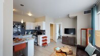 This single-floor space features an open-concept design that seamlessly connects the kitchen and living area. The kitchen is well-appointed with white cabinetry, stainless steel appliances including a gas range and microwave, and a dark slate tile backsplash that adds a contemporary contrast. A breakfast bar provides casual seating and reinforces the kitchen's connection to the living space. Light wood-look flooring extends throughout, contributing to the bright and airy feel of the interior. Neutral warm-toned walls keep the space feeling open and cohesive. The living area accommodates a media setup with a wall-mounted flat-screen TV positioned on a slatted media console, paired with a rustic round coffee table that adds a touch of warmth. A doorway visible from the main area leads to what appears to be a closet or additional storage, offering practical organization. The overall layout is compact yet efficient, making thoughtful use of every corner while maintaining a comfortable, livable atmosphere suitable for everyday use.