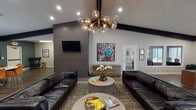 This single-story space is designed with a contemporary, art-forward sensibility that sets it apart from standard common areas. The main lounge is expansive and open, featuring soaring vaulted ceilings accented by bold black exposed beams that draw the eye upward. A striking multi-globe brass chandelier serves as a central focal point, casting warm light across rich wood-look flooring that runs throughout the space.

The lounge is furnished with sleek black leather sofas arranged around a round concrete-style coffee table, creating a comfortable yet sophisticated gathering area. A floor-to-ceiling grey tile accent wall houses a wall-mounted flat-screen television, while a gold credenza adds a luxurious touch nearby. Bold, large-scale pop-art prints and framed photography are displayed prominently on the walls, lending the space a gallery-like character.

Adjacent to the main lounge, a dining area features deep teal walls, a round white pedestal table, and vibrant orange velvet chairs, offering a colorful and intimate setting for group dining or meetings. Natural light filters in through multiple windows. A glimpse of a fitness area and a reception desk suggest this is a well-appointed community amenity space designed for both socializing and productivity.