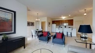 This single-floor residence offers a thoughtfully designed open-concept layout that seamlessly integrates the living, dining, and kitchen areas into one cohesive and functional space. The living area is anchored by a pair of navy blue armchairs accented with bold red throw pillows, arranged around a sleek glass-and-metal coffee table, creating an inviting focal point. Recessed lighting throughout keeps the space bright and airy, while neutral-toned carpeting adds warmth underfoot. The dining area is elegantly defined by a classic chandelier, providing a refined touch above a dark wood dining table and patterned upholstered chairs. Framed wall art adds a decorative element to the softly painted walls. The kitchen features rich, warm-toned cabinetry, a white refrigerator, and built-in microwave, all arranged for efficient use. A breakfast bar with seating connects the kitchen to the main living space, enhancing the open flow and making it ideal for casual dining or entertaining. The overall design balances practicality with comfort, utilizing clean lines and a neutral color palette accented with tasteful décor to create a polished, move-in-ready environment.