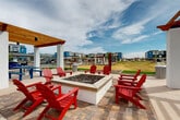 Located at 45760 W Village Parkway in Maricopa, Arizona, this outdoor community amenity space is thoughtfully designed to encourage social gathering and relaxation within a contemporary residential development. At the heart of the space sits a square fire pit surrounded by bold red Adirondack chairs, creating a welcoming focal point for evening gatherings. Two wood-topped pergolas with white structural columns provide shaded seating areas, each outfitted with blue picnic tables for casual outdoor dining or socializing. Flanking the central patio, stretches of lush artificial turf host suspended hammocks strung between upright wooden poles, offering a leisurely retreat. Desert-friendly landscaping with gravel ground cover and young trees frames the green zones, reflecting a low-maintenance aesthetic suited to the Arizona climate. Wide concrete walkways connect each area seamlessly, ensuring easy navigation throughout the space. Adjacent covered parking provides convenient, shaded vehicle storage for residents. The surrounding multi-story residential buildings in modern earth tones and bold accent colors are visible throughout, reinforcing the sense of a vibrant, well-planned community. Open skies above complete the inviting, resort-inspired atmosphere of this amenity area.