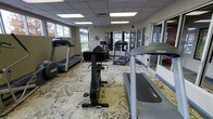 This fitness center offers a clean, functional environment designed to support a variety of workout routines. The space is laid out on a single floor and features a patterned carpet that adds a degree of comfort underfoot, distinguishing it from the typical hard-floored gym setting. A drop ceiling with recessed fluorescent panel lighting ensures consistent, even illumination throughout the room.

The facility is outfitted with multiple Precor-brand treadmills positioned along the perimeter, offering users views through the large windows that line the walls. Natural light filters in generously, creating a bright and welcoming atmosphere. A stationary upright bike occupies a central position on the floor, providing a low-impact cardiovascular option. Additional cardio and strength equipment is visible throughout, offering a well-rounded selection for users with varying fitness goals.

Large interior windows and glass partitions on one side of the room connect the fitness area visually to adjacent spaces, contributing to an open, airy feel. Wall-mounted mirrors help expand the sense of space while allowing users to monitor their form. The overall ambiance is practical and inviting, making this fitness center a comprehensive and accessible facility for daily exercise.