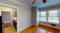 This single-floor residence combines classic architectural character with thoughtfully updated interiors. Warm hardwood floors run throughout, lending continuity and a timeless appeal to every room. The living areas are distinguished by soft blue-gray walls adorned with elegant white picture-frame moldings, crown molding along the ceilings, and ceiling fans that add both style and function. Natural light filters through multiple windows dressed with blinds, offering pleasant outdoor views and a bright, airy atmosphere. Baseboard heating is present throughout, ensuring year-round comfort.

The kitchen is efficiently designed and well-appointed, featuring dark espresso cabinetry, granite-style countertops, a stainless steel refrigerator, a gas range, an over-the-range microwave, and a classic white subway tile backsplash — a clean, functional space suited for everyday cooking. The bathroom retains a traditional feel with white subway tile surrounding a full soaking tub, a window for natural ventilation, and a wall-mounted mirror.

A long, narrow entryway corridor with hardwood flooring and crisp white baseboards leads to the main living spaces, creating a clear sense of entry and transition. Throughout the residence, the cohesive color palette, detailed millwork, and polished floors come together to create a warm, classic ambiance.