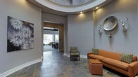 This well-appointed single-floor space makes a striking first impression with its grand rotunda entryway, framed by a curved tray ceiling with recessed lighting that draws the eye upward and creates an immediate sense of openness and architectural elegance. The flooring throughout the entry and main living areas is laid in rich, multi-toned slate tiles, adding texture and a grounded, earthy contrast to the smooth gray walls. The neutral gray palette flows seamlessly throughout, providing a clean and modern backdrop that complements the eclectic furnishings, including a warm cognac leather sofa, a patterned accent chair, and decorative wall-mounted art pieces including a sculptural deer head mount. A broad hallway extends from the rotunda, leading to a spacious gathering or dining area at the rear of the space, which is bathed in natural light streaming through large floor-to-ceiling windows and glass doors. This back room features dark-stained wood beam accents and ample seating, suggesting a comfortable lounge or event area. The overall ambiance is polished yet welcoming, blending contemporary architectural details with warm, curated décor to create a space that is both visually impressive and functionally versatile.