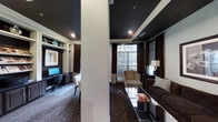 This single-floor space showcases a striking interior design defined by dramatic black ceilings contrasted against crisp white crown molding and structural columns, creating a bold and polished aesthetic throughout. The layout is divided into two distinct yet cohesive zones, each serving a different functional purpose.

The workstation area features a built-in desk with integrated shelving and cabinetry finished in white, providing a clean and organized workspace. Computer monitors are neatly housed within the setup, and decorative accents on the upper shelves add a touch of personality. Recessed lighting illuminates this area effectively, while rich chocolate-brown curtains frame the surroundings.

Adjacent to the workspace, the lounge area exudes a refined, comfortable atmosphere. A plush dark sofa, accent chairs, and a sleek glass coffee table are arranged around a large window that allows natural light to filter in, offering views of the surrounding greenery. A stylish table lamp and dark wood accent furniture further elevate the space.

Patterned carpeting runs throughout, tying both areas together cohesively. The overall ambiance is contemporary and sophisticated, balancing functionality with elegant design details.