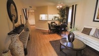 This single-floor residence showcases a well-designed open-concept layout that seamlessly connects the living, dining, and kitchen areas into one cohesive and inviting space. Rich hardwood-style flooring runs throughout the main living area, lending warmth and continuity to the interior. The living space is generously proportioned, offering ample room for comfortable furnishings, and is anchored by a plush area rug and tasteful décor accents including tall decorative vases with natural greenery. The dining area sits centrally within the open plan, featuring a round dark-finished table with matching chairs, perfectly suited for everyday meals or entertaining. A ceiling fan with integrated lighting provides both comfort and illumination overhead. Adjacent to the dining area, the kitchen is accessible via a raised breakfast bar pass-through, offering a practical and social connection between the cooking and living spaces. White cabinetry and under-cabinet lighting give the kitchen a clean, bright appearance. Natural light filters in through windows dressed with horizontal blinds and dark curtain panels, creating a warm yet refined ambiance. A hallway leading from the main living area provides access to additional rooms, completing this functional and well-appointed single-level layout.