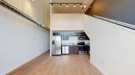 This two-story loft-style residence in Memphis, Tennessee offers a distinctive open-concept layout defined by dramatic soaring ceilings on the main level. The living and kitchen areas share a bright, open floor plan finished with light wood-style flooring throughout. The kitchen is tucked beneath the staircase overhang and is outfitted with dark cabinetry, granite-style countertops, and a full suite of stainless steel appliances, including a side-by-side refrigerator with ice and water dispenser, electric range, built-in microwave, and dishwasher. Exposed ductwork and track lighting add an industrial loft character to the space.

The upper level serves as the bedroom, accessible via a staircase and separated from the main level by a glass-panel railing that allows visual openness between floors. The bedroom is carpeted and lit with track lighting, offering a cozy contrast to the open living space below.

The full bathroom features a combination tub and shower, chrome fixtures, a toilet, and wood-style flooring, maintaining a clean and functional aesthetic consistent with the rest of the unit. Overall, the space blends industrial loft design elements with practical, modern finishes.