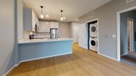 This single-floor unit showcases a contemporary design with a neutral gray and white color palette carried throughout. The kitchen is outfitted with white shaker-style cabinetry, white quartz countertops, a classic subway tile backsplash, and a full suite of stainless steel appliances, including a refrigerator, electric range, and over-the-range microwave. Pendant lighting above the kitchen island adds a modern touch, while light hardwood flooring extends seamlessly into the open living area. A stacked washer and dryer unit is conveniently tucked into a dedicated closet just off the main living space.

The primary bedroom features hardwood flooring and a large window that provides views of the surrounding urban streetscape, filling the room with natural light. The secondary bedroom is finished with plush gray carpeting and a narrower vertical window that also overlooks the exterior. The full bathroom includes large-format gray tile flooring, a white tub and shower combination with a curved curtain rod, and brushed nickel fixtures, maintaining a clean, cohesive aesthetic. Overall, the unit presents a modern, functional layout with consistent finishes suited for comfortable urban living.