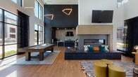 This contemporary amenity lounge is a thoughtfully designed communal space that blends bold aesthetics with versatile functionality. Soaring double-height ceilings create an expansive, airy atmosphere throughout, while large floor-to-ceiling windows flood the interior with abundant natural light and offer views of the outdoor pool area and surrounding streetscape.

The main gathering area serves as the focal point, anchored by a dramatic built-in linear fireplace framed in dark penny tile and wood-toned stone cladding, with a tiled bench seat adorned with colorful accent pillows. Adjacent to the fireplace, a stylish lounge seating area features a caramel leather sectional, a bold black-and-white striped rug, and an eye-catching copper wire wall art installation that spans the full height of the wall.

A full-size billiards table is centrally positioned beneath striking geometric pendant lights, complementing the dark paneled accent wall with bold architectural millwork detailing. The wet bar area is appointed with dark granite countertops, white shaker cabinetry, a black matte sink, a built-in microwave, and a bold star-patterned tile backsplash, with a mounted television above.

A dedicated co-working nook offers a printer station alongside a sculptural open wood bookshelf partition, completing this richly appointed community space.