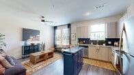 This thoughtfully laid-out one-bedroom, one-bathroom unit offers a seamless blend of modern design and functional living across a single floor. The open-concept kitchen and living area serves as the heart of the home, featuring light wood cabinetry, granite countertops, a bold navy blue kitchen island with a stainless steel top, and a full suite of stainless steel appliances including a refrigerator, range, microwave, and dishwasher. A striking dark slate tile backsplash adds contrast and visual interest to the kitchen. Wood-style plank flooring runs throughout the main living space, complementing the bright, neutral color palette. Large windows dressed with navy curtains flood the living area with natural light and offer views of the surrounding neighborhood. A ceiling fan adds comfort to the space. The living area also features a unique built-in display niche, adding architectural character to the clean white walls. The bedroom is carpeted for warmth and comfort, offering a walk-in closet and elevated transom windows that bring in additional natural light. The bathroom includes a full tub-shower combination with white tile surround, maintaining the unit's clean, contemporary aesthetic throughout.