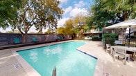Located in the desirable Houston Heights area, this outdoor amenity space at 2121 Tannehill Dr offers a well-appointed pool and deck environment designed for relaxation and recreation. The centerpiece is a generously sized swimming pool with clear turquoise water, flanked by a combination of concrete pool surround and a multi-level wood deck that provides ample space for lounging. The deck features teal-cushioned lounge chairs paired with small side tables, creating comfortable seating areas for residents to unwind. A striking mature tree rises directly through the deck, offering natural shade and a charming focal point that enhances the outdoor atmosphere. The space is enclosed by a horizontal-plank gray privacy fence, lending a modern aesthetic while creating a secluded retreat. String lights are draped around the tree, adding a warm ambiance for evening gatherings. Additional seating is available near a covered umbrella table on the pool deck, providing options for shaded outdoor dining or casual conversation. The surrounding canopy of mature trees filters natural light throughout the day, contributing to a peaceful, park-like setting. This outdoor space serves as a communal hub, combining practical amenities with an inviting, well-landscaped environment in the heart of Houston.