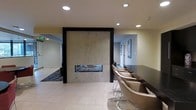 This single-floor space presents a sleek, contemporary design centered around a dramatic floor-to-ceiling marble fireplace surround framed in dark espresso wood. The linear gas fireplace serves as the room's focal point, adding warmth and visual interest to the open layout. Light-toned ceramic tile flooring extends throughout the main area, contributing to the bright, clean aesthetic complemented by recessed ceiling lighting. To one side, a row of modern bar-height chairs in neutral tones lines a dark counter surface, creating a casual seating area ideal for socializing or working. Framed artwork adorns the adjacent wall, adding a curated touch to the environment. An open corridor visible beyond the fireplace suggests additional connected spaces, with patterned carpeting marking the transition. The overall ambiance is polished and inviting, blending warm materials like marble and dark wood accents with a neutral color palette to create a space that feels both functional and refined, well-suited for lounging or gathering.