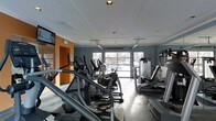 This fitness center is a purpose-built, single-floor space designed to support a full cardio and exercise regimen. The room is outfitted with a robust lineup of cardio equipment, including elliptical trainers, stair climbers, and additional aerobic machines arranged in orderly rows to maximize floor space and ease of movement between stations. The interior features a polished concrete floor, which lends a clean and durable foundation suited for high-traffic use. Recessed ceiling lights provide consistent, even illumination throughout the space, complementing the natural light that pours in through large, floor-to-ceiling glass garage-style doors along the far wall. These doors offer views to the exterior and contribute to an open, airy atmosphere. The walls are painted in a cool blue-gray tone, accented by a warm orange accent wall near the entrance, creating a visually dynamic environment. A wall-mounted television is positioned for viewing during workouts. Full-length mirrors on one side of the room enhance the sense of space and allow users to monitor their form. The overall ambiance is modern, functional, and inviting, making it a well-considered facility for daily fitness activity.