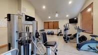 This dedicated fitness facility is thoughtfully designed to support a full range of workout routines. The space is organized across a single floor and features vaulted ceilings in key areas, contributing to an open and airy atmosphere. Recessed lighting throughout ensures the space is well-illuminated, complemented by natural light from multiple wood-trimmed windows dressed with warm-toned blinds.

The main workout area is generously appointed with a multi-station cable strength machine, an adjustable workout bench, and a dumbbell rack, providing ample options for resistance training. A workout reference chart is posted for user guidance. The cardio section is equally well-stocked, featuring multiple elliptical trainers, a stationary upright bike, and a dedicated treadmill room with two SportsArt treadmills side by side.

Multiple wall-mounted flat-screen televisions are strategically positioned throughout the space, allowing users to stay entertained during their workouts. Stability balls and additional accessories are also on hand for functional training. The flooring transitions between polished concrete in the main workout zone and carpeting in the treadmill room, defining each area functionally. Clean, neutral walls and warm wood trim details lend the facility a polished yet welcoming character.