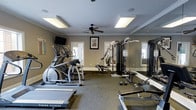 Located at 2605 Elms Plantation Blvd in Charleston, South Carolina, this single-floor fitness center offers a well-appointed space designed to support a comprehensive workout routine. The room features neutral, warm-toned walls accented by crisp crown molding, giving the space a polished and welcoming atmosphere. Durable, high-traction carpet flooring covers the workout area, providing stability and comfort during exercise. Natural light filters in through multiple windows, complemented by recessed skylight-style ceiling panels that ensure the space remains bright and energizing throughout the day. A ceiling fan circulates air to keep the environment comfortable during workouts. The fitness center is equipped with a variety of machines, including an elliptical trainer, a treadmill, and a multi-station cable and weight machine, catering to both cardio and strength training needs. A free-weight station with dumbbells and a flat bench offers additional versatility. Full-length mirrors line one wall, enhancing the sense of space and allowing users to monitor their form. A wall-mounted television adds an entertainment option, making workouts more engaging. This fitness center provides a clean, functional, and well-maintained environment for residents seeking convenient access to quality exercise facilities.