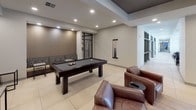 This single-floor Lobby & Residents Lounge combines a welcoming entrance with a well-appointed social space. The lobby greets visitors through a striking floor-to-ceiling glass and steel entryway, flooded with natural light from the large arched transom windows. A circular pendant light fixture anchors the ceiling, casting a warm glow over the large-format tiled floors. A concierge-style desk and an adjacent conference area provide functional workspace just off the main entry.

Transitioning into the residents lounge, the space opens up into a generously sized recreation and gathering area. A full-size billiards table takes center stage, surrounded by rich brown leather club chairs that invite casual conversation and relaxation. A wall-mounted flat-screen television and a sleek linear gas fireplace serve as a focal point on one wall, creating a warm and inviting ambiance. Decorative tiled accent panels add visual interest and texture to the space.

A kitchenette equipped with cabinetry, a coffee station, and prep space supports resident gatherings with ease. Recessed lighting throughout provides even, comfortable illumination, while the tray ceiling detail in the lounge area adds architectural depth. Large glass doors and windows maintain a visual connection to the outdoors, contributing to an open, airy atmosphere throughout.