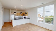 Located at 2820 West Broad Street in Richmond, Virginia, this one-bedroom, one-bathroom unit offers a clean, modern aesthetic throughout. Light hardwood flooring flows seamlessly across the entire space, creating a warm and cohesive feel. The open-concept kitchen and living area form the heart of the unit, where a peninsula with dark granite countertops provides both prep space and a natural divider. The kitchen is well-appointed with light maple cabinetry, white lower cabinets, stainless steel appliances including a full-size refrigerator, electric range, and built-in microwave, complemented by a classic white tile backsplash. Three statement pendant lights hang above the peninsula, adding a contemporary touch. Large windows in both the living area and bedroom flood the space with natural light and offer elevated views of the surrounding Richmond cityscape. The bedroom is a comfortable, well-proportioned retreat featuring a ceiling fan and a louvered closet door. The full bathroom includes a soaking tub and shower framed by bold blue tile, warm wood cabinetry, dark granite countertops, and a full vanity mirror, striking a stylish contrast against the white walls.