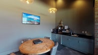 Located at 4125 Hayden Lofts Pl in Dublin, Ohio, this single-floor commercial space combines modern design with a warm, professional atmosphere. The interior is finished in deep, muted tones — most notably a rich slate-green on the walls — which lend the space a contemporary and refined character throughout.

The most prominently visible area features a comfortable meeting or lounge nook centered around a solid wood round table with upholstered chairs, ideal for small gatherings or client consultations. A wall-mounted flat-screen display adds a functional media element to the space, while a large pendant light and an additional decorative ceiling fixture provide warm, focused illumination that enhances the intimate ambiance.

Adjacent to the seating area is a well-appointed kitchenette or refreshment station, outfitted with lower cabinetry finished in the same muted green tone as the walls, dark stone countertops, and a utility sink. The counter space accommodates various appliances and accessories, making it a practical and stylish amenity for occupants and guests alike.

The flooring features dark tile, contributing to the cohesive, upscale aesthetic carried throughout the space. The overall design is thoughtfully curated to feel both functional and welcoming, making this Dublin location well-suited for professional or boutique commercial use.