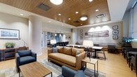 Located at 201 S 25th Street in Philadelphia, PA, this single-floor space offers a well-appointed, open-concept layout designed for both comfort and functionality. The main gathering area features a warm, inviting atmosphere anchored by a rich leather sofa, modular seating, and a mix of lounge and dining furniture arranged to encourage both relaxed socializing and focused activities. Distinctive wood-slat ceiling panels add warmth and texture overhead, complemented by a combination of recessed lighting and statement pendant and flush-mount fixtures that create a well-lit, layered ambiance throughout. Structural columns with clean, curved profiles contribute to the architectural character of the space without interrupting the open flow. Toward the rear, a fully equipped kitchen area is visible, showcasing navy cabinetry, stainless steel appliances, and pendant lighting — ideal for group gatherings or events. On the opposite end, a dedicated workspace or dining zone features booth-style seating and task lighting, offering a more private setting within the larger open floor plan. Light hardwood flooring and neutral-toned area rugs ground the space, while framed artwork and decorative accents lend a polished, contemporary finish. Overall, the space balances modern design with practical versatility.