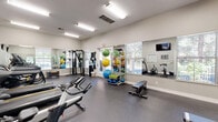 Located at 7300 Parkwood Cir in Dublin, California, this fitness center offers a thoughtfully equipped exercise space designed to support a wide range of workout routines. The single-floor layout provides an open, functional environment finished with durable rubber flooring and bright white walls that contribute to a clean, energizing atmosphere. Flush-mounted fluorescent lighting keeps the space well-lit throughout, while multiple windows with blinds allow natural light to filter in and offer views of the surrounding greenery outdoors. Large wall-mounted mirrors run along several walls, enhancing the sense of openness and allowing users to monitor their form during exercise. The cardio zone features multiple Matrix treadmills, cycling bikes, and additional cardio machines, each positioned for comfortable use. Multiple wall-mounted flat-screen TVs are positioned throughout the room for entertainment during workouts. The strength and functional training area is well-stocked with a cable machine, adjustable weight bench, dumbbells, plyo boxes, and an extensive collection of stability and medicine balls neatly organized on a rack. A glass-paneled door provides direct access to an outdoor area, further connecting the space to its surroundings. Informational signage is posted near the entrance, adding a practical touch to this comprehensive and versatile fitness facility.