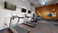 This fitness facility is thoughtfully organized across a single floor, offering distinct zones tailored to a variety of workout styles. The main exercise area is generously proportioned, featuring high ceilings with exposed ductwork and track lighting that creates an open, industrial-inspired ambiance. The space is anchored by a row of Matrix-brand treadmills and elliptical trainers, each equipped with individual monitors, while multiple wall-mounted flat-screen televisions provide additional entertainment throughout. A bold orange accent wall adds a dynamic visual element to the cardio section.

The strength training zone features a cable machine, adjustable bench, spin bike, and a rack of dumbbells, complemented by wall mirrors that enhance the sense of space. Colorful modular carpet tiles cover the main exercise floor, providing both comfort and durability. A well-stocked accessory rack holds stability balls and resistance tools near the accent wall.

Adjacent to the main gym, a separate studio room features light wood-look flooring, soaring walls, track lighting, and a wall-mounted television, making it suitable for group fitness or stretching classes. A secondary enclosed room with the same flooring houses a freestanding boxing bag, ideal for martial arts or combat training.