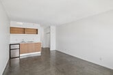 Located on Garland Avenue in the heart of Los Angeles, CA, this compact studio unit offers a functional and efficiently designed single-floor layout. The open living area features polished concrete floors throughout, lending an industrial-modern aesthetic that pairs well with the crisp white walls and textured ceiling. Large windows along one side of the living area flood the space with natural light and frame elevated views of the surrounding Los Angeles cityscape, with a wall-mounted HVAC unit conveniently positioned beneath them.

The integrated kitchenette is neatly tucked along one wall and includes warm wood-toned lower cabinetry with sleek metal hardware, a white countertop, a stainless steel sink with a single-handle faucet, an electric cooktop, and open upper shelving in a contrasting orange wood finish — adding a pop of color and accessible storage.

The bathroom is clean and straightforward, featuring light beige ceramic tile flooring, white subway tile walls around the tub-shower combination, a wood-toned vanity with an integrated sink, a large mirror, and overhead lighting. The overall design of the space is minimal and functional, making it well-suited for urban living in a central Los Angeles location.