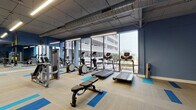 Located at 17105 Gull Blvd N in North Redington Beach, Florida, this fitness center offers a well-equipped, single-floor space designed for a comprehensive workout experience. The facility features an industrial-inspired aesthetic, with exposed ductwork running along the ceiling and sleek linear lighting that illuminates the space effectively.

Floor-to-ceiling windows line one wall, flooding the gym with natural light and providing views of the surrounding coastal environment, including palm trees and neighboring structures. The windows create an open, airy atmosphere that enhances the workout environment.

The cardio zone is positioned along the windowed wall and includes multiple treadmills, elliptical machines, and stationary bikes, allowing users to exercise while enjoying the outdoor views. A dedicated strength and functional training area features a Precor cable machine, TRX suspension training straps, and a rack of medicine balls, providing versatile options for resistance and functional workouts. Adjustable benches are distributed throughout the floor, supporting a variety of exercises.

The flooring consists of neutral-toned carpet tiles accented with bold blue and teal insets, adding visual interest while defining workout zones. Deep blue accent walls complement the coastal color palette. A mirrored wall in the functional training area expands the sense of space and allows for form monitoring during exercise.