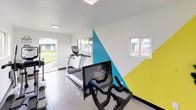 The Cardio Lab is a dedicated fitness space designed to deliver a focused cardiovascular workout experience. The room is finished with durable concrete flooring and crisp white walls, punctuated by a striking geometric mural rendered in teal, yellow, and white — adding an energetic, modern aesthetic that motivates movement. Recessed and flush-mounted lighting keeps the space bright, while multiple windows along the walls invite ample natural light and offer views of the surrounding green outdoor areas. A glass door provides direct access to an outdoor walkway, further enhancing the open, airy feel of the room. The equipment lineup includes a True elliptical trainer, a treadmill, and a Peloton stationary bike equipped with a large interactive touchscreen display for live and on-demand fitness classes. A potted fiddle-leaf fig plant adds a touch of greenery to the corner, softening the industrial elements. Gym rules are posted near the entrance, reflecting a well-maintained and community-oriented environment. Compact yet thoughtfully arranged, this single-level cardio lab provides everything needed for an effective aerobic training session.