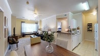 This single-floor residence offers a thoughtfully designed open-concept layout that effortlessly connects the dining, living, and kitchen areas into one cohesive and inviting space. The dining area features rich hardwood flooring and flows naturally into the carpeted living room, which is anchored by a ceiling fan and a large window dressed with layered curtains that allow soft, natural light to fill the room. Crown molding adds a refined architectural detail throughout the common areas, lending the space a polished, classic character. The kitchen is appointed with crisp white cabinetry, dark granite countertops, and a breakfast bar that provides both additional seating and a natural boundary between the kitchen and the living space. Tile flooring in the kitchen area offers a clean, practical transition from the hardwood and carpet of the adjoining rooms. A hallway visible from the main living area leads to the bedroom, offering a sense of depth and privacy within the floor plan. The overall ambiance is warm and comfortable, with a neutral color palette of whites and soft yellows creating a bright, airy atmosphere. The cohesive design and functional layout make this a highly livable and visually appealing space.