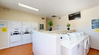 Located at 330 Waymore Dr in El Paso, Texas, this community laundry facility is a clean, efficiently designed single-room space built to serve residents with convenience and comfort. The room is finished with warm terracotta tile flooring and neutral cream-toned walls, creating a bright and welcoming atmosphere. Fluorescent overhead lighting ensures consistent illumination throughout.

The facility is well-stocked with multiple front-loading washing machines and a set of stacked washer-dryer units lining the walls, all white and uniformly arranged for easy access. A central white folding counter runs through the heart of the space, providing ample surface area for sorting and folding laundry. Decorative potted fiddle-leaf fig plants add a refreshing touch of greenery to the utilitarian environment.

A wall-mounted flat-screen television is positioned in a corner, offering entertainment while residents wait. Large windows along one wall flood the room with abundant natural light and offer pleasant views of an outdoor pool area, enhancing the overall ambiance. A long folding table is positioned near the windows, providing additional workspace.

The space is accessed through a charming paneled door and includes clearly posted instructional signage throughout. The layout is thoughtfully organized to maximize functionality while maintaining a tidy, orderly appearance.