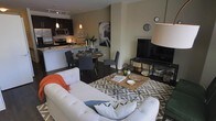 This single-floor residence offers a thoughtfully designed open-concept layout that blends living, dining, and kitchen spaces into one cohesive and inviting environment. The living area serves as the heart of the home, anchored by a plush white tufted sofa accented with colorful throw pillows and a bold geometric-patterned area rug. A wooden coffee table with glass legs sits at the center, complementing the entertainment setup nearby. The dining area features a round wooden table surrounded by contemporary gray chairs, creating an intimate setting ideal for everyday meals or entertaining guests. The kitchen is sleek and modern, outfitted with dark cabinetry, stainless steel appliances including a microwave and refrigerator, and a light-toned counter with bar seating. Pendant lighting adds warm, focused illumination over the kitchen and dining zones, while track lighting runs along the ceiling to brighten the overall space. Dark hardwood-style flooring flows consistently throughout, providing a rich contrast to the neutral warm-toned walls. Decorative artwork and thoughtfully placed accents add personality and visual interest to the space. The overall ambiance is modern, warm, and welcoming, with a design aesthetic that balances comfort and contemporary style.