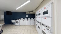 This shared laundry facility is a practical, well-organized space designed to serve a residential community. The room is finished with light-colored ceramic tile flooring throughout, providing a clean and easy-to-maintain surface. A bold navy blue accent wall adds a touch of color to one side of the room, contrasting with the neutral gray and white tones elsewhere.

The facility is equipped with a row of front-loading and top-loading washing machines, each clearly numbered for easy identification, along with multiple stacked dryer units lining the walls. The machines are managed through a CSC payment system, with posted instructions, service information, and laundry tips displayed prominently for user convenience.

A wall-mounted utility sink offers additional functionality, while a large granite-topped folding table provides ample space for sorting and folding laundry. A waste bin is conveniently located nearby. The room is brightly lit by overhead fluorescent strip lighting, ensuring excellent visibility at any time of day.

Ventilation is provided via ceiling-mounted HVAC units, maintaining a comfortable environment. A standard exterior door with a window allows for natural light to enter and provides easy access to the outdoors. The overall layout is spacious and uncluttered, making for an efficient and user-friendly laundry experience.