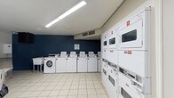 This shared laundry facility is a practical, well-organized space designed to serve a residential community. The room is finished with light-colored ceramic tile flooring throughout, providing a clean and easy-to-maintain surface. A bold navy blue accent wall adds a touch of color to one side of the room, contrasting with the neutral gray and white tones elsewhere.

The facility is equipped with a row of front-loading and top-loading washing machines, each clearly numbered for easy identification, along with multiple stacked dryer units lining the walls. The machines are managed through a CSC payment system, with posted instructions, service information, and laundry tips displayed prominently for user convenience.

A wall-mounted utility sink offers additional functionality, while a large granite-topped folding table provides ample space for sorting and folding laundry. A waste bin is conveniently located nearby. The room is brightly lit by overhead fluorescent strip lighting, ensuring excellent visibility at any time of day.

Ventilation is provided via ceiling-mounted HVAC units, maintaining a comfortable environment. A standard exterior door with a window allows for natural light to enter and provides easy access to the outdoors. The overall layout is spacious and uncluttered, making for an efficient and user-friendly laundry experience.