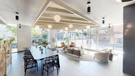 Located at 517 N Shipley Street in Wilmington, Delaware, this thoughtfully designed communal space offers a sophisticated and welcoming atmosphere across a single open floor. The layout is defined by striking exposed wooden beam ceilings that add warmth and architectural character, complemented by an array of globe pendant lights that provide soft, ambient illumination throughout.

Floor-to-ceiling windows line the perimeter, flooding the interior with abundant natural light and offering open views of the surrounding streetscape and greenery. Large-format marble-toned tile flooring anchors the space with an upscale, polished aesthetic.

The lounge area features a curated selection of contemporary furnishings, including plush sofas, accent chairs, and sculptural coffee tables arranged around a sleek fireplace with a wall-mounted display above, creating an inviting focal point. A dedicated dining and collaborative work zone offers a long communal table with modern seating, ideal for group gatherings or focused work sessions.

A distinct booth-style seating area, accented with richly upholstered navy banquettes and decorative gold lattice partitions, provides a more intimate setting. Lush potted plants are positioned throughout, enhancing the connection to nature and contributing to a vibrant, refreshing ambiance. A beverage station is also accessible within the space, adding everyday convenience.