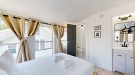 Located in the heart of downtown Saint Petersburg, Florida, this single-floor room offers a well-designed, self-contained layout ideal for comfortable short- or long-term stays. The space is anchored by a queen-sized bed positioned beneath large wraparound windows dressed with neutral grommet curtains, allowing abundant natural light while framing views of the surrounding cityscape and downtown skyline. The warm wood-look flooring throughout adds a clean, contemporary feel that contrasts nicely with the crisp white walls and a decorative light blue patterned accent wall. A wall-mounted flat-screen TV and a compact mini-fridge provide in-room convenience, while a gold-finish rolling garment rack offers practical storage. Framed artwork adds a personalized touch to the décor. The en-suite bathroom is accessible through a dark panel door and features a toilet, vanity with sink and mirror, and a tiled shower with curtain — all finished in a clean, minimalist style. A second door provides access to the exterior, enhancing the room's sense of openness and accessibility. The space is thoughtfully appointed with fresh towels and a small bedside table, making it fully functional. Situated steps from Saint Petersburg's vibrant downtown core, the location offers easy access to dining, entertainment, and waterfront amenities.