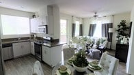 This single-floor home features a thoughtfully designed open-concept layout that seamlessly connects the kitchen, dining, and living areas into one cohesive living space. The kitchen is outfitted with crisp white cabinetry, dark countertops, and stainless steel appliances, including a gas range and built-in microwave, offering both functionality and a clean aesthetic. A window above the sink allows natural light to filter in, brightening the workspace. The dining area sits just adjacent to the kitchen, centered around a glass-top table elegantly set with woven placemats and a lush floral centerpiece, adding a fresh, organic touch. Wood-look flooring runs throughout, lending warmth and continuity across the open floor plan. The living room receives generous natural light through large windows dressed with two-tone gray and white curtains, complemented by a ceiling fan overhead. Neutral-toned furnishings accented with green throw pillows echo the botanical elements found throughout the space, creating a cohesive, inviting ambiance. A sliding glass door provides access to an outdoor balcony, enhancing the sense of openness and connection to the exterior.