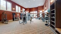 Located at 14909 Health Center Drive in Bowie, Maryland, this single-floor health and fitness center offers a comprehensive wellness environment designed for a wide range of activities. The main fitness floor is a generously proportioned space featuring soaring ceilings and an abundance of natural light streaming through large, multi-pane windows set high along the deep crimson walls. The gym is well-appointed with a full array of strength training machines, a dumbbell rack, ellipticals, treadmills, exercise balls, and wall-mounted mirrors that amplify the sense of space. Accent lighting and a mounted flat-screen TV complete the workout environment. Adjacent to the main gym, a dedicated studio features rich hardwood flooring, deep teal accent walls, floor-to-ceiling mirrors, globe pendant lighting, and exercise mats, making it ideal for yoga, stretching, or group classes. A children's activity area—labeled "Fun Station"—offers built-in shelving, a chalkboard wall, patterned carpeting, and seating, accessible through glass-panel doors. The facility opens to an outdoor courtyard with a fenced swimming pool, a decorative water fountain feature, and a pergola with classical columns, providing a refined outdoor retreat surrounded by mature trees.