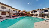 Located at 1829 E Morten Ave in Phoenix, Arizona, this outdoor communal space centers around a large, generously sized swimming pool with crystal-clear turquoise water. The pool area is framed by a two-story residential building with a classic mid-century architectural style, featuring stucco exteriors, decorative lattice screens, and private balconies with wrought iron railings that overlook the courtyard below.

The pool deck is finished with a smooth concrete surround and is well-appointed with resort-inspired outdoor furniture, including cushioned chaise lounges and sectional seating in bold navy and orange tones. A shaded dining area with a red umbrella and bistro-style table and chairs provides a comfortable space for outdoor gatherings. Towering palm trees and desert-adapted landscaping add a lush, tropical ambiance that complements the warm Arizona climate.

The courtyard layout wraps the pool on multiple sides, creating an enclosed, community-centered atmosphere ideal for relaxation and socializing. Pool access points include steps and a stainless steel entry rail. The space benefits from abundant natural sunlight year-round, with an open sky that showcases the characteristic clear blue skies of the Phoenix region. Overall, this outdoor area delivers a well-maintained, inviting desert oasis atmosphere.